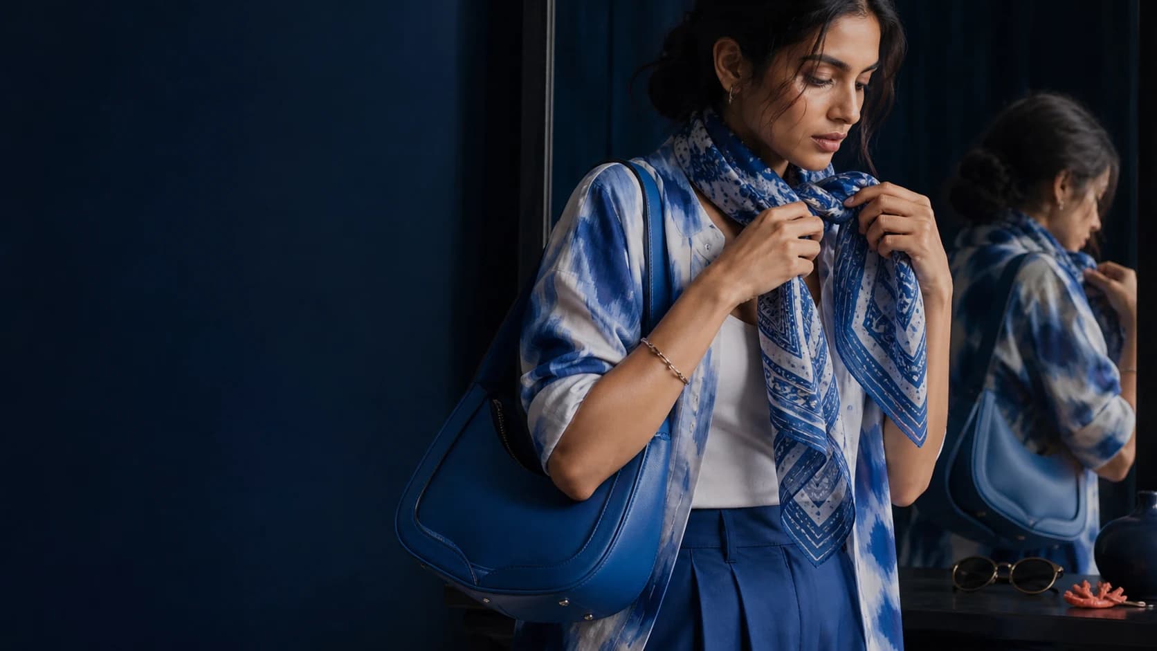A model styling a blue scarf and shoulder bag beside a studio mirror